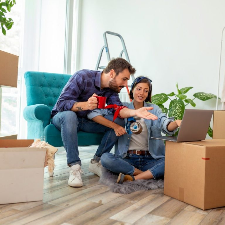 Vermietung Junges Paar in einem Wohnzimmer, das Laptop verwendet und Umzugskisten um sich hat.