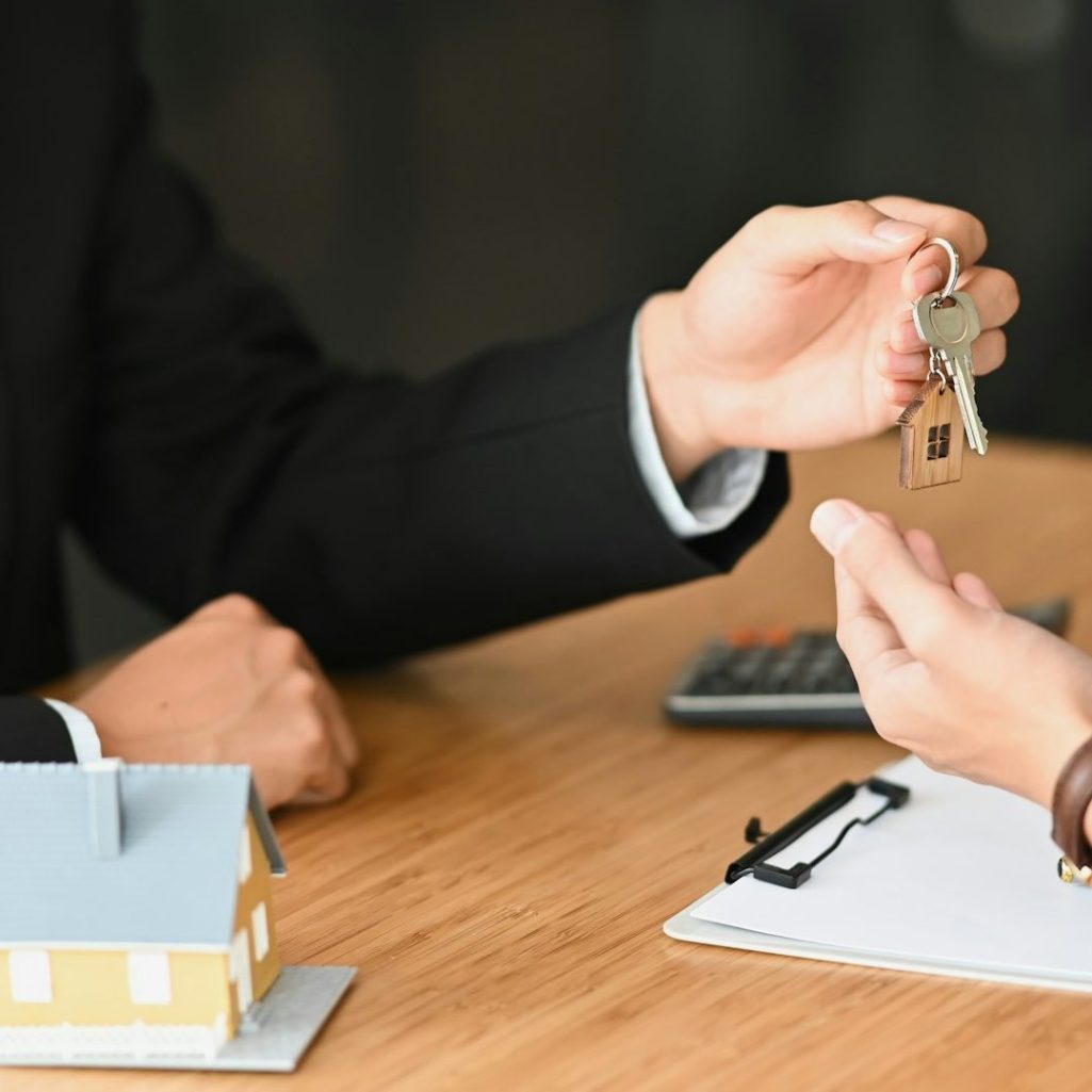 Wohnungsübergabe Ein Schlüsselübergabe von einer Person an eine andere, im Hintergrund ein Modellhaus.
